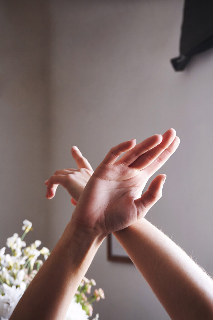 Marintia Bustos's hands delicately intertwine showcasing the intricate details of human anatomy against a serene backdrop of a room with soft lighting and a vase of white flowers.